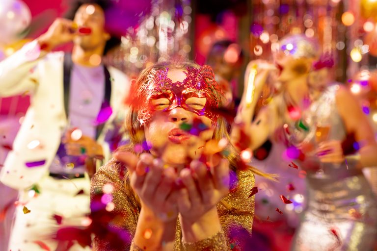 Happy caucasian woman in mask blowing glitter to camera at a party in a nightclub. Fun, going out, celebrating and party concept